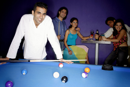 Young man standing at pool table, people in the background, all looking at camera