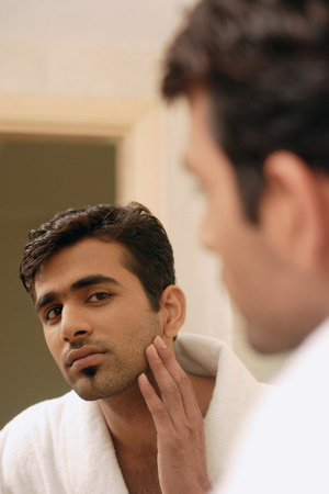 Man wearing bathrobe, looking in mirror, touching his faceの写真素材