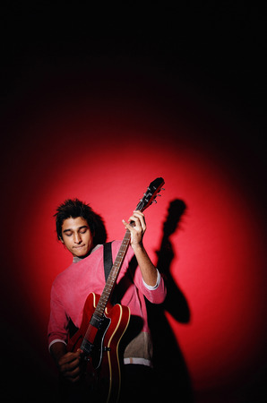 Young man playing guitar, standing against red backgroundの写真素材