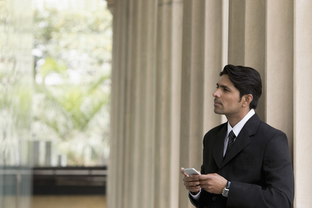 Businessman in front of columns looking up from mobile phoneの写真素材