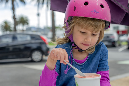 Young girl in pink outfit and pink helmet riding a kick-scooter sitting at the outside cafe table with icecreamの写真素材