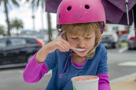 Young girl in pink outfit and pink helmet riding a kick-scooter sitting at the outside cafe table with icecreamの写真素材