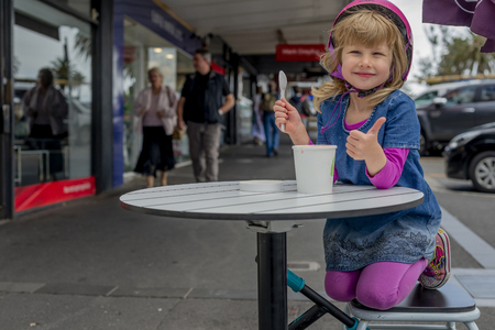 Young girl in pink outfit and pink helmet riding a kick-scooter sitting at the outside cafe table with icecreamの写真素材