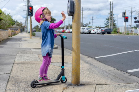 Young girl in pink outfit and pink helmet riding a kick-scooter pressing button at pedestriam crossingの写真素材
