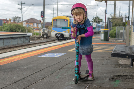Young girl in pink outfit and pink helmet riding a kick-scooter waiting on a platform at train stationの写真素材