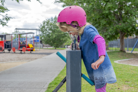 Young girl in pink outfit and pink helmet riding a kick-scooter in the park having a drink from fountainの写真素材