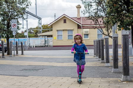 Young girl in pink outfit and pink helmet riding a kick-scooter on sidewalkの写真素材