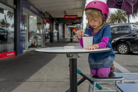 Young girl in pink outfit and pink helmet riding a kick-scooter sitting at the outside cafe table with icecreamの写真素材