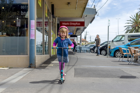 Young girl in pink outfit and pink helmet riding a kick-scooter on sidewalkの写真素材