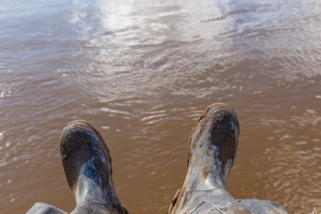 Feet of a traveler or wild nature explorer in black dirty boots sitting on the bank of the riverの写真素材