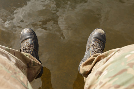 Low section of person above water. Men's legs in camouflage pants and old boots dangle above the...
