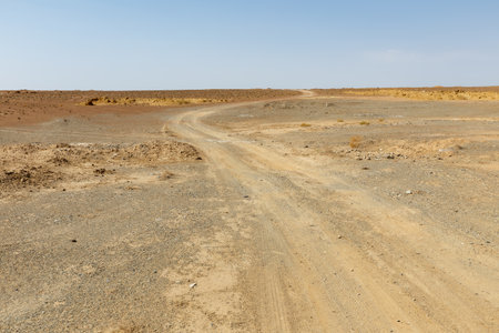 A winding dirt road cuts through a dry, barren landscape under a clear blue sky, showcasing the isolation of the area.の写真素材