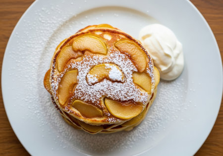 An overhead view of a delicious stack of apple pancakes, dusted with powdered sugar. This homemade breakfast is served on a white plate with whipped cream.の素材