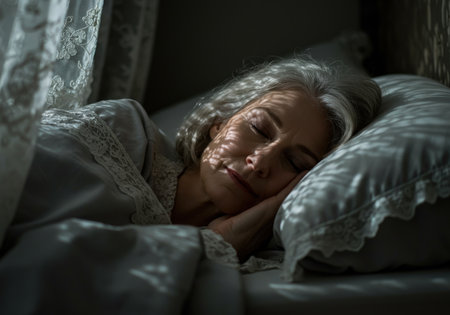 Peaceful senior woman deep in dream sleep illuminated by intricate morning sun shadows on her face. Her restful slumber is emphasized by the detailed lace curtains.の素材