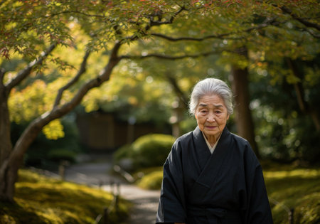 A calm and graceful senior Asian woman with gray hair, wearing a traditional black kimono. She stands in a beautiful Japanese park during autumn.の素材