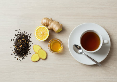 Cup of tea is surrounded by fresh ginger, lemon slices, honey, and loose black tea on a wooden surfaceの素材
