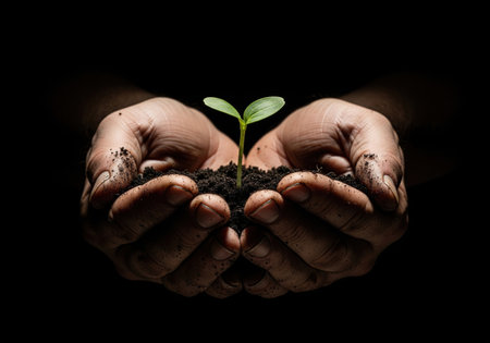 Farmer's hands holding a green sprout with soil, a concept of new life, growth and hope isolated on a dark black background.の素材