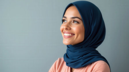 A joyful woman with a hijab smiles warmly while standing against a gray backdrop, exuding confidence and positivity.の素材