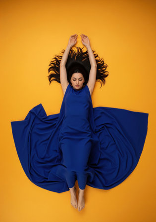 Beautiful woman in a blue dress lying on a vibrant yellow background. A minimalist top view fashion shot with strong color contrast.の素材