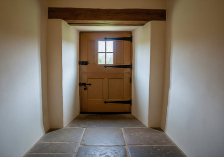Rustic wooden stable door with small window in white hallway interior with stone floor, featuring black iron hinges and oak beam.の素材