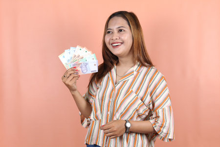 A happy young woman is wearing striped dress with brown hair and holding cash money in Indonesian rupiah isolated by peach backgroundの写真素材