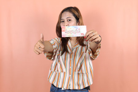 Portrait of happy Indonesian Muslim woman holding rupiah money isolated over peach background. Currency of 75000 Rupiah the 75th of Independence Day of Indonesia.の写真素材