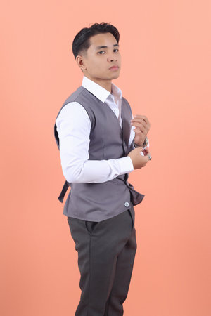 Handsome young Asian businessman wearing a suit vest is seen from the side adjusting the buttons of his sleeves confidently in peach background.の写真素材