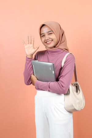 Beautiful young Asian woman holding a laptop while carrying a bag with her other hand waving while closing her eyes and smiling to show her teeth.の写真素材