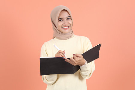 Smiling beautiful muslim Asian woman in cream sweater holding notebook pen and writing looking at...