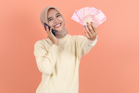 Excited beautiful Asian Muslim woman in cream sweater holding cash money in Indonesian rupiah...