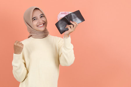 Excited young Asian muslim woman in cream sweater holding wallet full of money rupiah banknotes of...