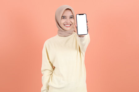 Excited beautiful young Asian Muslim woman wearing hijab and sweater showing smartphone with empty...