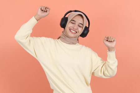 Beautiful young Asian woman in a cream sweater raises arms and dances to favorite music, forgot about all problems isolated over peach background have funの写真素材