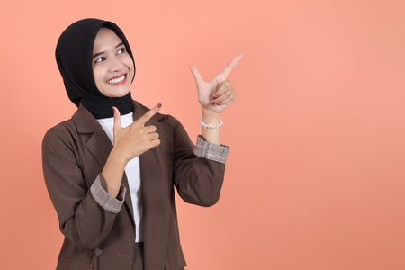 Excited beautiful Asian woman in brown suit and hijab pointing finger aside to copy space. People religious lifestyle conceptの写真素材