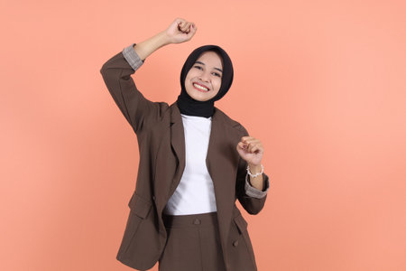 Enthusiastic muslim asian woman rejoicing, say yes, looking happy and celebrating victory, champion dance, fist pump gesture, wearing brown suit standing over peach backgroundの写真素材