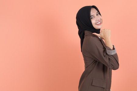 Excited beautiful Asian woman in brown suit and hijab pointing finger aside to copy space. People religious lifestyle conceptの写真素材