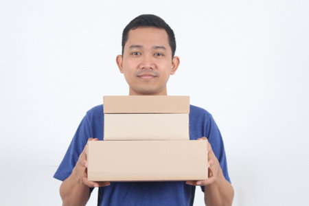 Image of a happy young Asian delivery man in blue standing with parcel post box isolated over white background.の写真素材