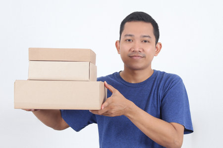 Image of a happy young Asian delivery man in blue standing with parcel post box isolated over white background.の写真素材