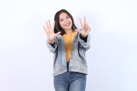 Young beautiful Asian woman wearing blue jacket yellow t-shirt showing number 8 and pointing up with finger number eight isolated on white backgroundの写真素材