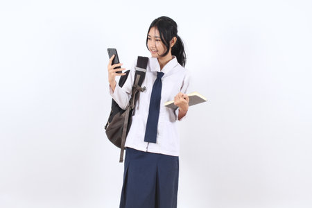 Asian teenage girl wearing a school uniform is looking at her cell phone. She is holding a book and a backpackの写真素材