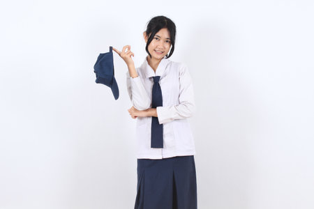 Asian teenage schoolgirl in traditional uniform holding and looking at a navy cap with a soft smile. Standing in front of a white background, the girl shows a casual and thoughtful expression.の写真素材