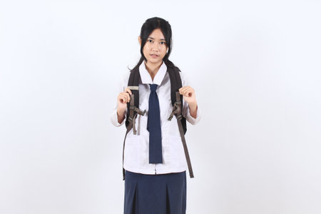 Happy Asian teenage girl in school uniform carrying a backpack and smiling, standing against a white...