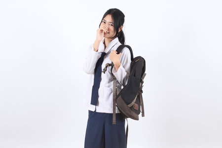 Happy Asian teenage girl in school uniform carrying a backpack and smiling, standing against a white...