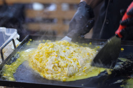 Freshly made cheesy corn with melted mozzarella being prepared by a cook on a flat-top grill. A...