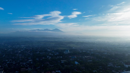 Breathtaking aerial view of a misty city in Indonesia at sunrise. A majestic mountain range and mosque rise above the tranquil morning landscape.の写真素材