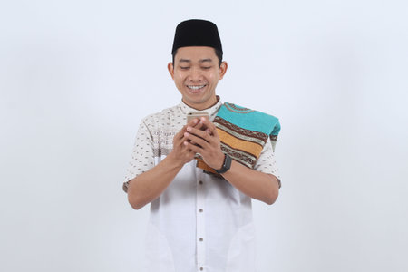Young happy Asian Muslim man in traditional clothes smiles while texting greetings on his mobile phone for Eid al-Fitr or Ramadan celebration.の写真素材