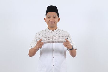 Confident Indonesian man in Islamic clothing smiling and pointing fingers at the viewer. Concept for recruitment, invitation, Ramadan, or Eid al-Fitr.の写真素材