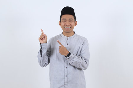 Cheerful Indonesian man in Muslim attire presenting an offer, pointing towards empty space for your ad. Perfect for Ramadan or Eid promotions.の写真素材