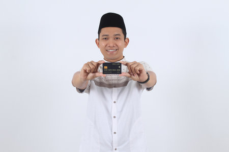 Smiling young Indonesian man in traditional Islamic clothes shows his bank card. Concept for e-commerce, digital payment, online shopping, and finance.の写真素材
