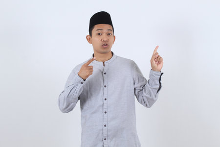 Young Asian Muslim man in traditional Islamic clothing points up, explaining a concept or having an idea. Isolated on white background for Ramadan or Eid.の写真素材